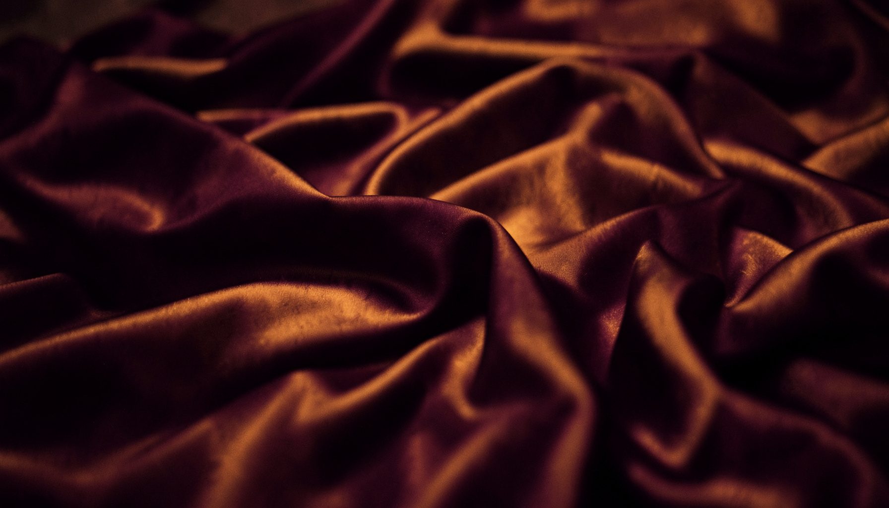 Soft duotone photograph of rumpled silk sheets in deep plum and warm amber tones, intimate and moody