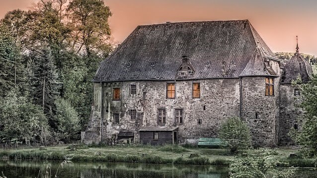 Weathered stone building with a steep dark roof and glowing window lights, surrounded by trees and a pond, warm sunset scene