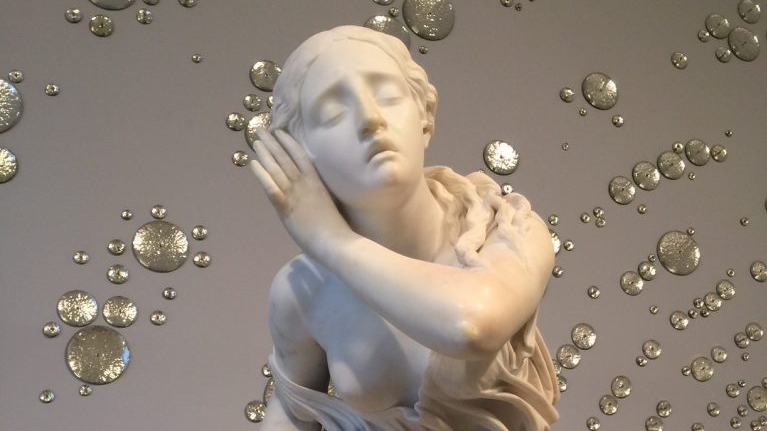 White marble statue of a reclining woman with closed eyes, set against a wall decorated with reflective silver disc ornaments